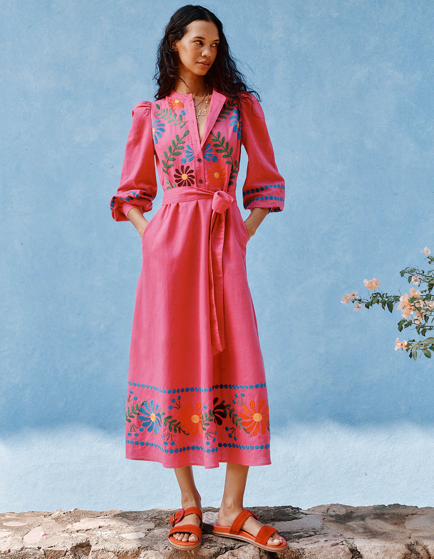 Frau in einem rosa Kleid mit Blumenstickerei vor blauem Himmel.