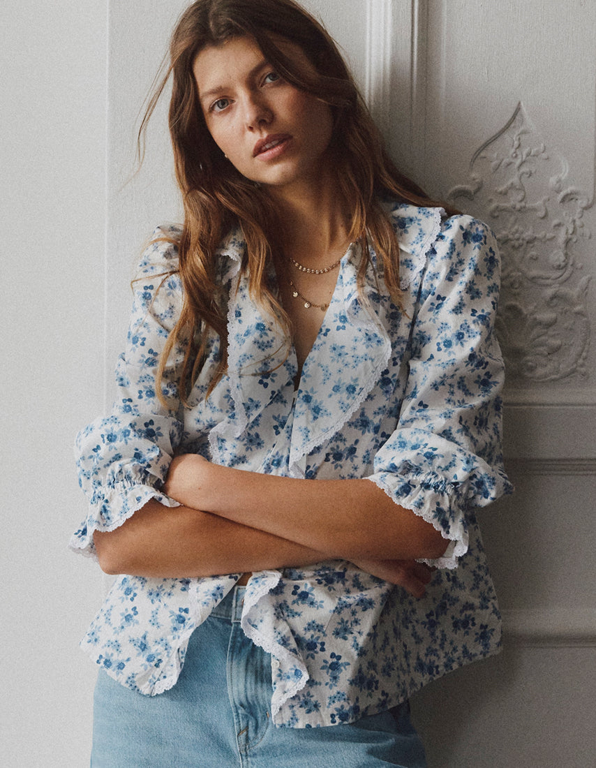 Frau in blauer Bluse mit Blumenmuster und Jeans vor einer hellen Wand.